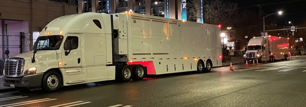 Nighttime street scene with a large white semi-truck in the foreground and another truck in the background near a lit building with banners.