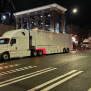 Nighttime street scene with a large white semi-truck in the foreground and another truck in the background near a lit building with banners.