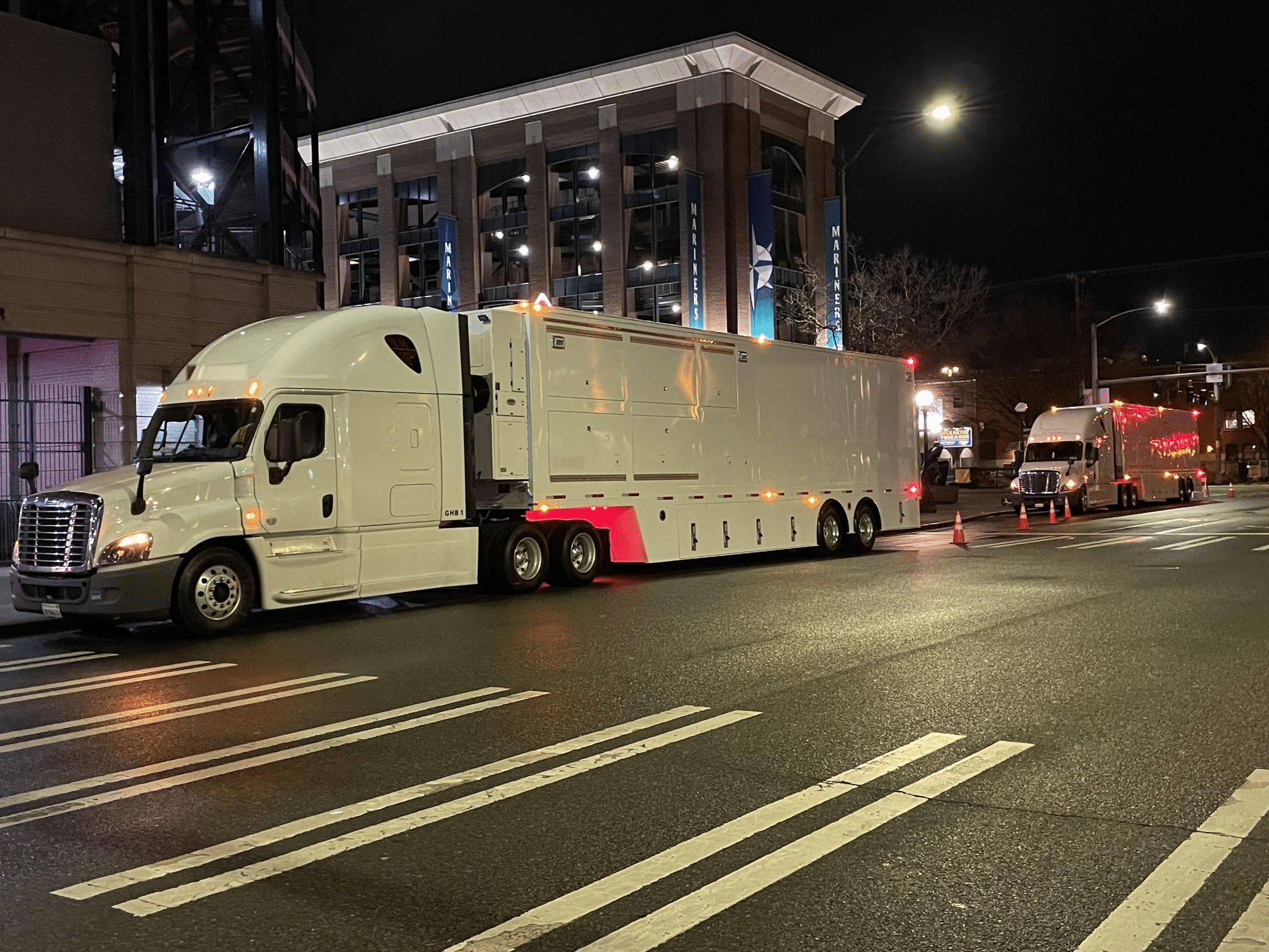 Nighttime street scene with a large white semi-truck in the foreground and another truck in the background near a lit building with banners.